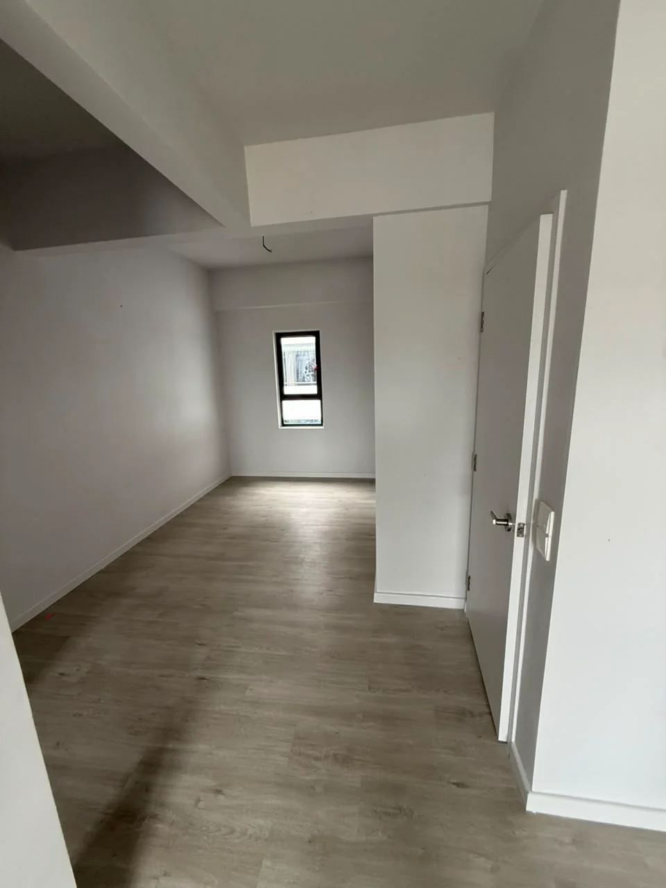 Room features: light wood laminate flooring, white painted walls, single window, plain ceiling, door, corner layout; style: modern.